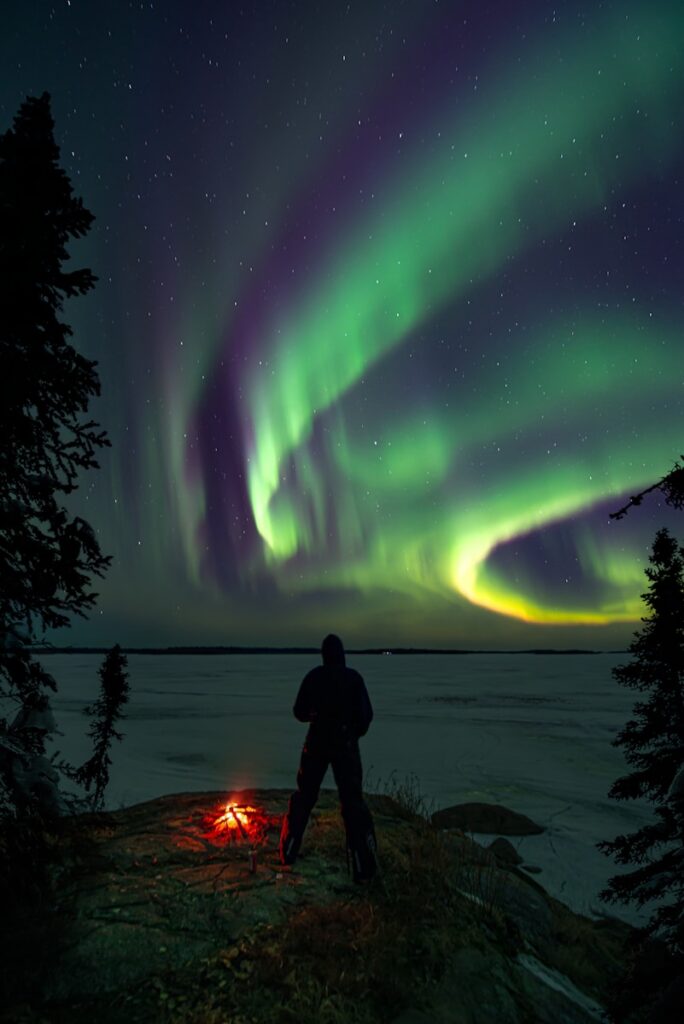 a man standing next to a campfire under an aurora bore